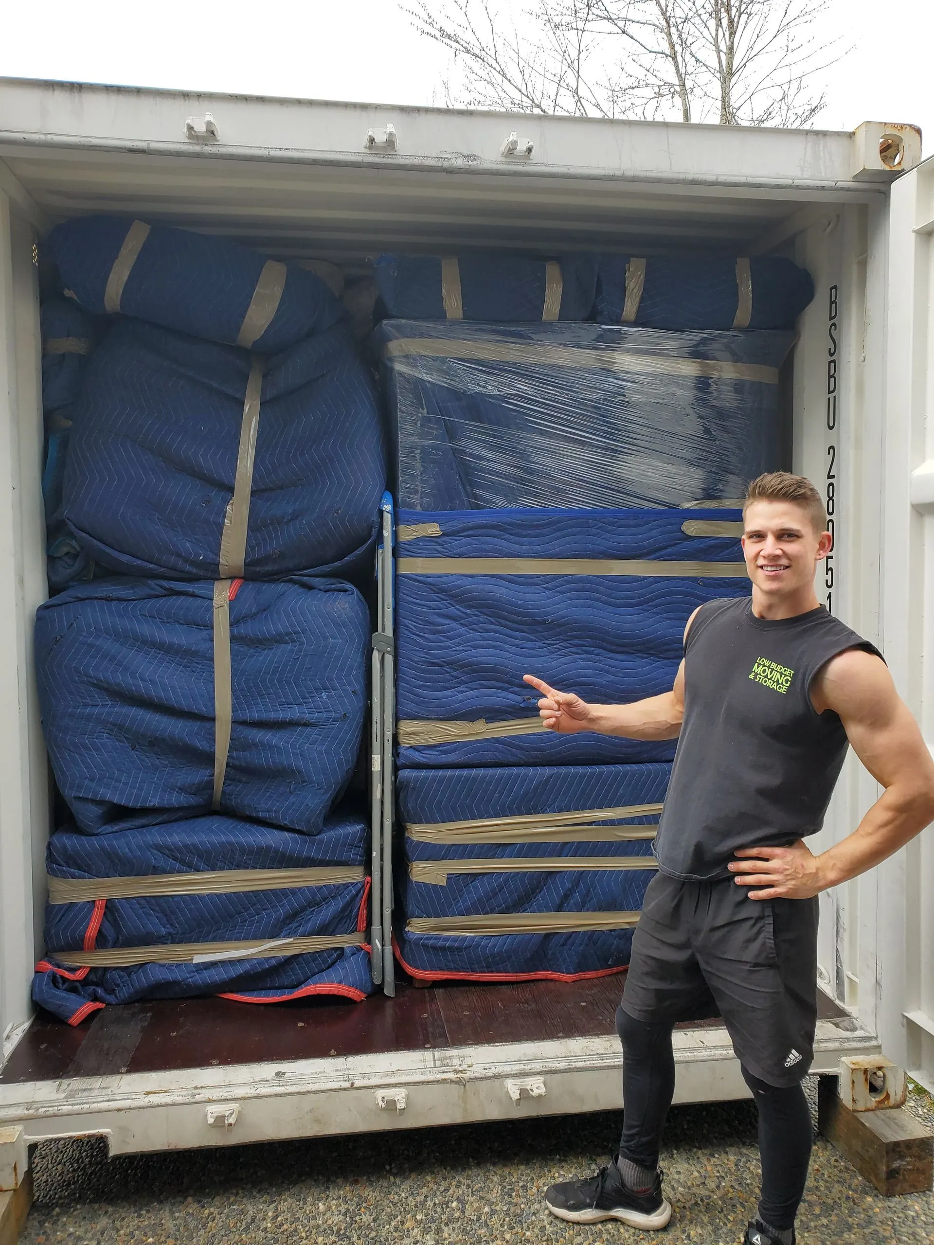 Low Budget Moving crew member beside furniture wrapped in blue moving blankets in a container