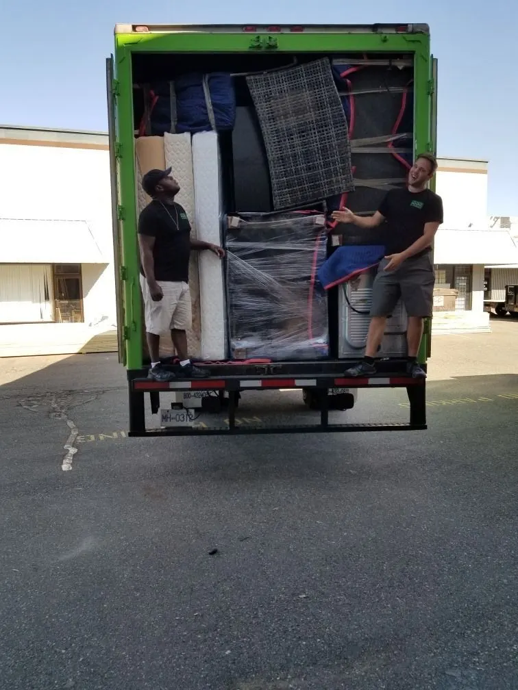 Two movers loading blanket-wrapped furniture into the back of a green moving truck