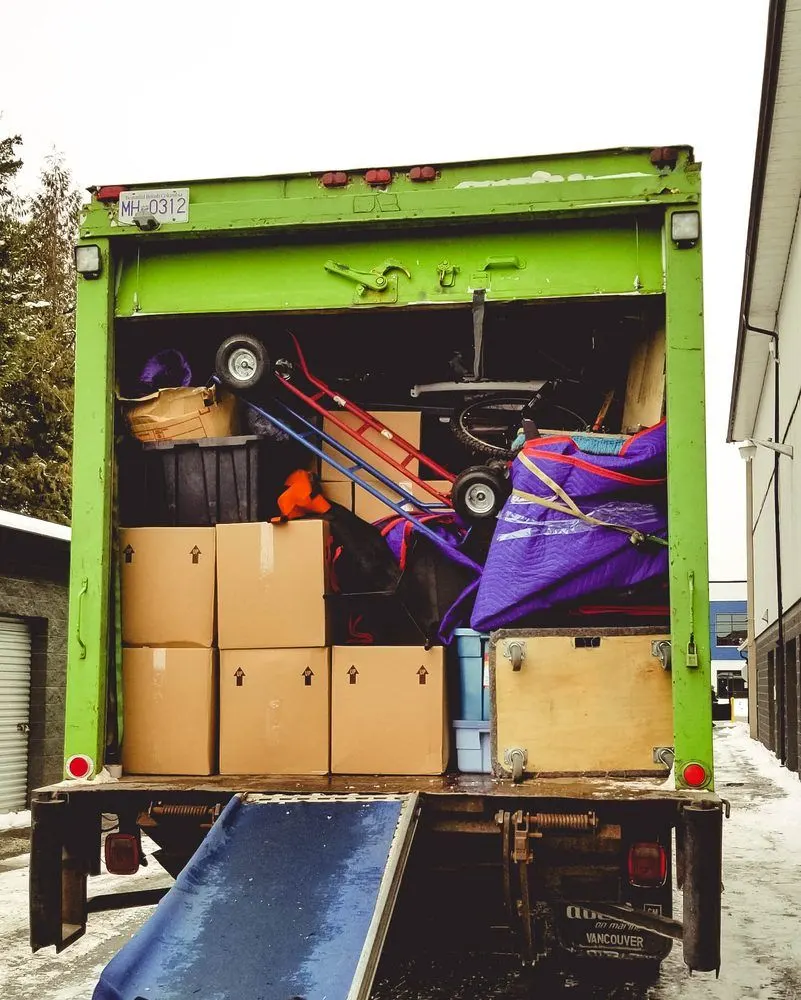 Back of a green moving truck packed with boxes, a dolly and furniture, ramp lowered
