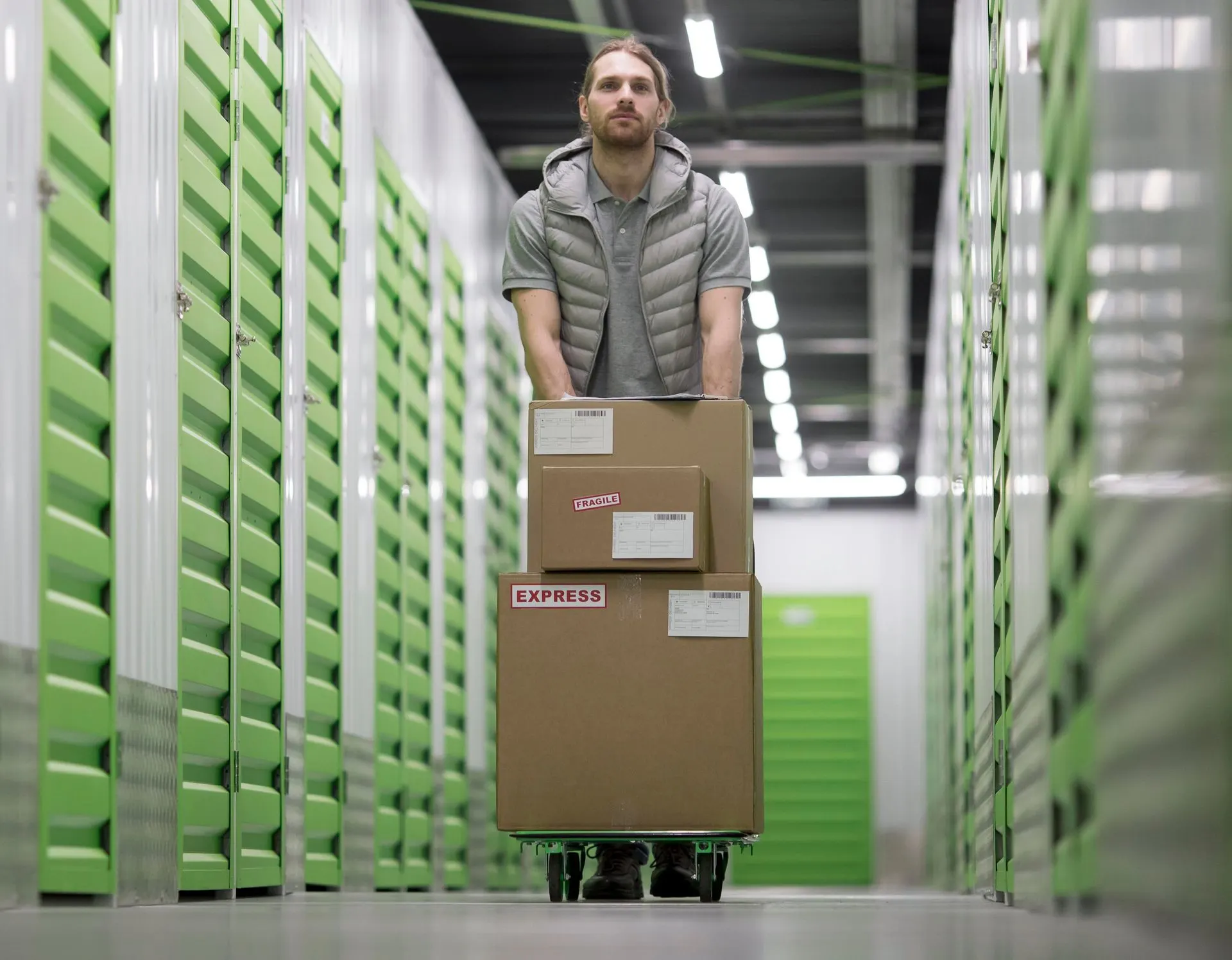 Man pushing a hand truck of boxes down a bright storage facility aisle with green doors