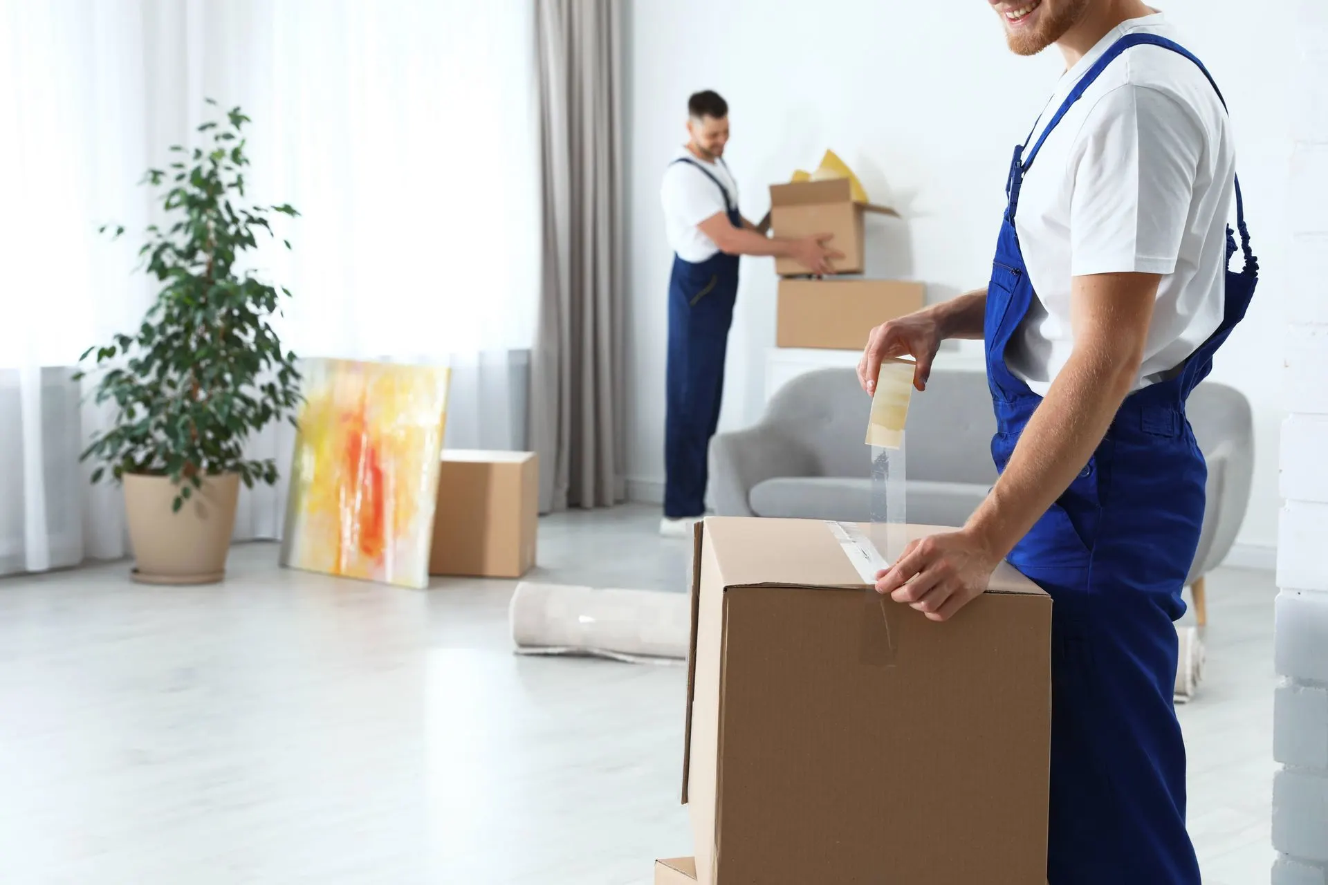 Mover in blue overalls taping a cardboard box while a coworker carries boxes in a bright home