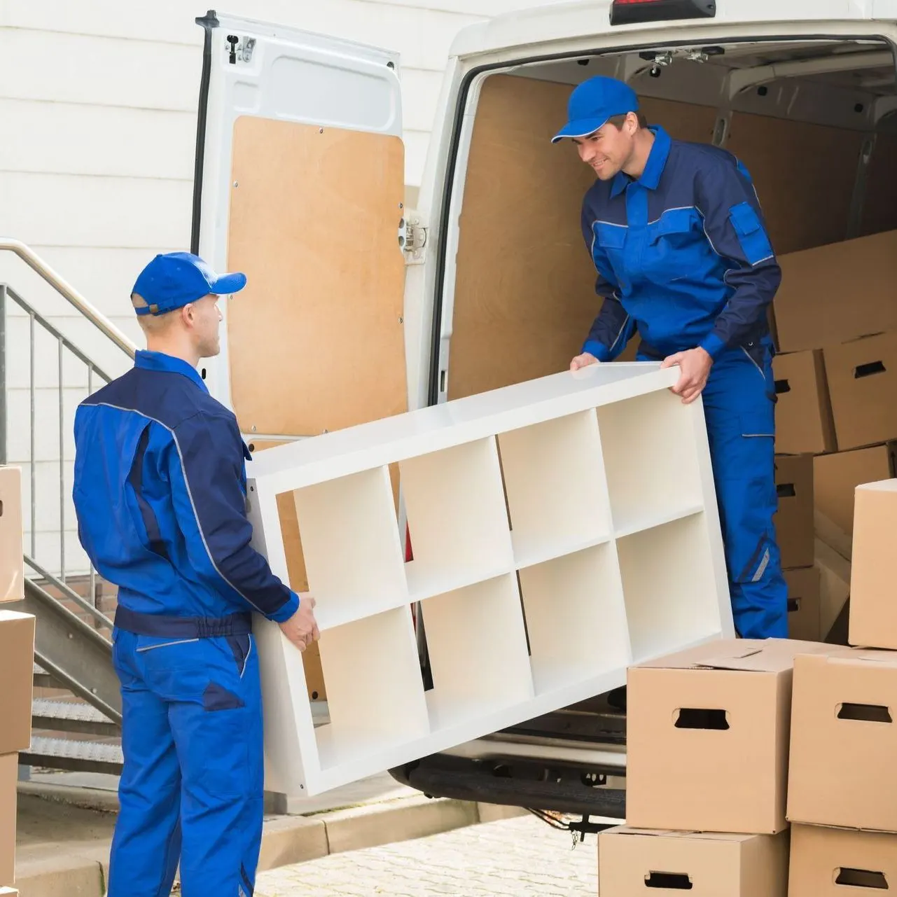 Two movers in blue uniforms lifting a white shelving unit out of a moving van